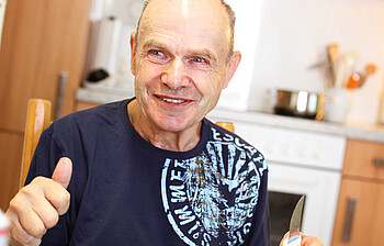 Beinahe kahlköpfiger Mann mittleren Alters, der ein dunkelblaues t-Shirt trägt und wahrscheinlich gerade am Essen ist, da er in seiner linken Hand ein Messer hält. Er sitzt auf einem gemütlichen Holzstuhl an einem Tisch und lächelt dabei. 