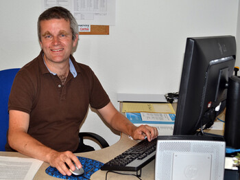 Ein Herr mittleren Alters sitzt an einem Schreibtisch vor einem Computer. Er trägt ein braunes Polo Shirt und hält seine rechte Hand auf der Computermaus. 