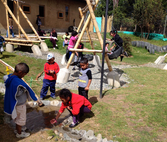 Ein Spielplatz im Freien, auf dem zahlreiche Kinder spielen. Manche sind beim Schaukeln, andere spielen mit dem Wasser. 