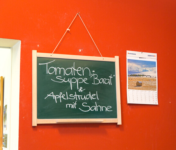 An einer roten Wand hängt eine holzumrahmte grüne Tafel, auf der das Mittagessen des Caritas-Cafés für den jeweiligen Tag mit weißer Kreide steht. 