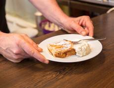 Ein Apfelstrudel mit Sahne wird auf einem weißen kleinen Teller serviert. Auf dem Teller befindet sich auf eine Gabel. 