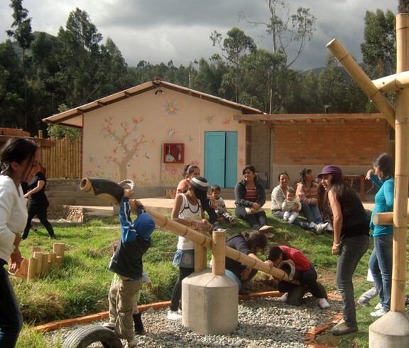 Viele Kinder, die im Freien spielen. Schauplatz ist ein Spielplatz. Im Hintergrund sitzen einige Mütter mit ihren Kindern.