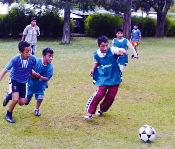 Eine Gruppe junger Buben, die beim Fußballspielen sind. Ein Junge will gerade den Ball schießen, die anderen laufen ihm hinterher.