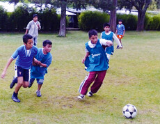 Eine Gruppe junger Buben, die beim Fußballspielen sind. Ein Junge will gerade den Ball schießen, die anderen laufen ihm hinterher.