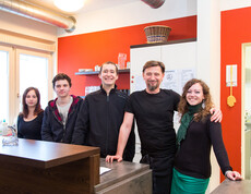 Die Mitarbeiter des Caritas-Cafés stehen im Café zu einem Gruppenfoto zusammen. Es sind fünf Personen, zwei Frauen und drei Männer. Sie befinden sich hinter der Theke. Die Wand im Hintergrund ist orange. Auch steht hinter ihnen ein großer weißer Schrank. 