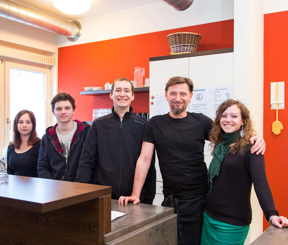 Die Mitarbeiter des Caritas-Cafés stehen im Café zu einem Gruppenfoto zusammen. Es sind fünf Personen, zwei Frauen und drei Männer. Sie befinden sich hinter der Theke. Die Wand im Hintergrund ist orange. Auch steht hinter ihnen ein großer weißer Schrank. 