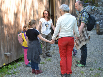 Drei Kinder stehen mit drei Erwachsenen in einem Kreis im Freien zusammen. Dabei halten sich alle am Arm.