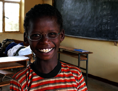 Ein kleiner Junge sitzt in einer Schulklasse. Er trägt eine Brille mit hellen Gläsern und strahlt. 