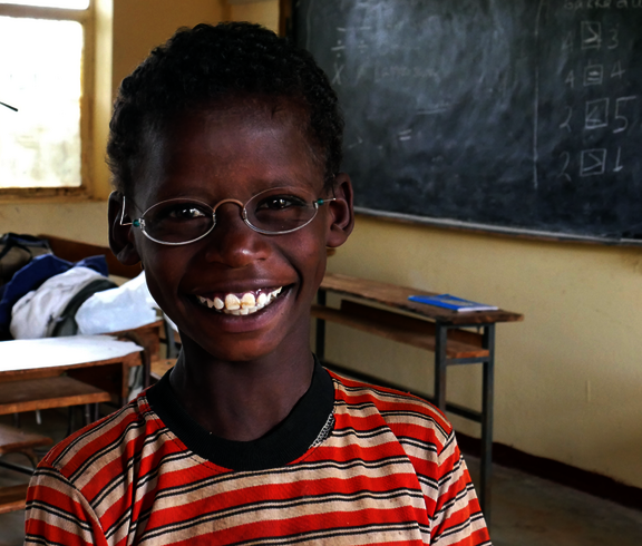 Ein kleiner Junge sitzt in einer Schulklasse. Er trägt eine Brille mit hellen Gläsern und strahlt. 