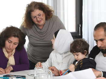 Eine Frau mittleren Alters lernt mit einer Flüchtlingsfamilie. Auf dem Tisch befinden sich Blätter, Bücher und Stifte. Die Frau steht dabei hinter der Familie. Die Familie sitzt am Tisch.