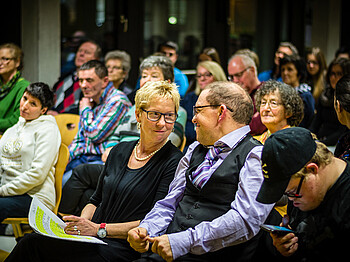 Viele Menschen sitzen im Publikum bei einer Präsentation. Im Vordergrund sieht man eine Dame, die einen Herrn anlächelt.