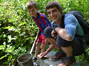 Ein junger Erwachsener und ein Bub sind in der Natur. Der junge Bursche hat einen Fußballtrikot an und hält einen Stock in der Hand. Der Erwachsene trägt einen Rucksack und steht auf einem großen Stein. Beide haben kutze Haare. Der Hintergrund schücken grüne Gräser. 