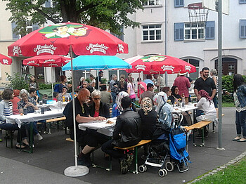 Eine Feier im Freien mit vielen Menschen. Sie sitzen auf Holzbänken und essen.