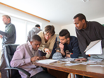 Flüchtlinge aus Afrika und dem nahen Osten lernen Deutsch. Sie sitzen gespannt und konzentriert an einem Tisch und blicken auf ihre Unterlagen. Eine Lehrerin hilft ihnen dabei den Stoff besser aufzunehmen und steht für jede Frage offen. 