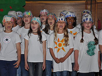 Um die zehn Mädchen im Volksschulalter stehen zusammen und singen. Sie tragen alle lange Haare, weiße T-Shirts und selbstgebastelte Kopfbedekungen. 