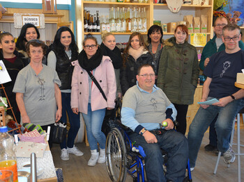 Menschen mit und ohne Behinderung stehen zu einem Gruppenfoto zusammen. Ein Mann sitzt in einem Rollstuhl. Sowohl junge als auch ältere Personen sind vertreten. Sie sind in einem Einkaufsladen. 