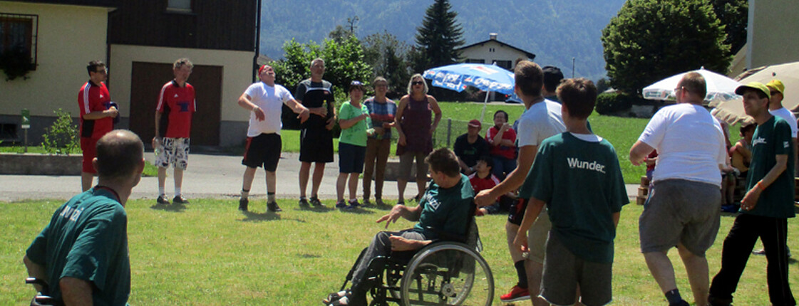 Mehrere Klienten der Werkstätten spielen auf einem grünen Rasen Fußball. Zwei von Ihnen sitzen dabei in ihrem Rollstuhl.