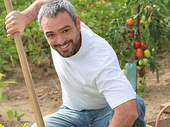 Ein Asylwerber mittleren Alters wird durch die Nachbarschaftshilfe beschäftigt. Er ist in einem Garten, hält einen Rechen in der Hand und lächelt in die Kamera. Er hat ein schlichtes weißes t-Shirt und eine Jeans an. Im Hintergrund sind Tomatenstöcke. 