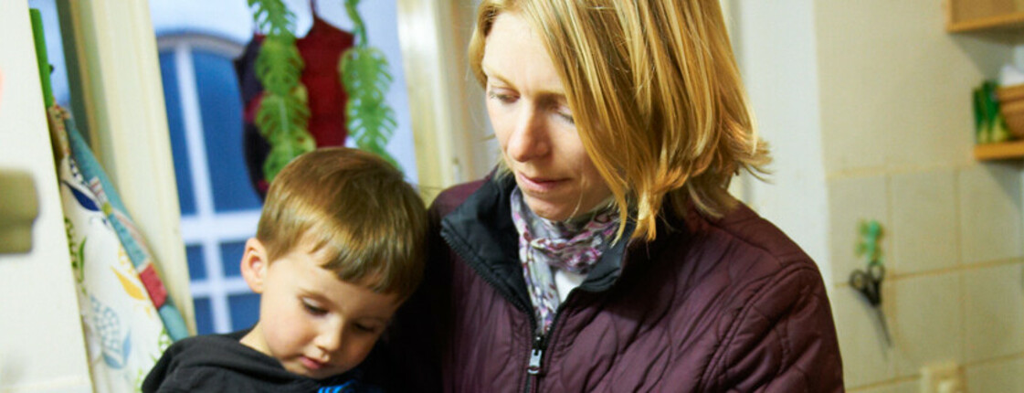 Eine Mutter mit einem kleinen Bub befindet sich in einer engen, älteren Küche. Der Bub hat sie dabei auf dem Arm.  Die Mutter ist allem Anschein nach mit der Situation überfordert. 