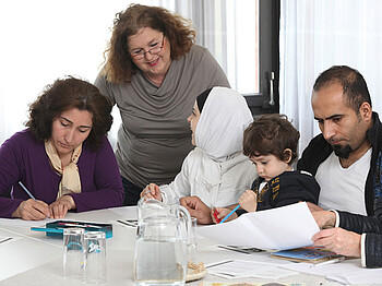 Eine Frau mittleren Alters lernt mit einer Flüchtlingsfamilie. Auf dem Tisch befinden sich Blätter, Bücher und Stifte. Die Frau steht dabei hinter der Familie. Die Familie sitzt am Tisch.