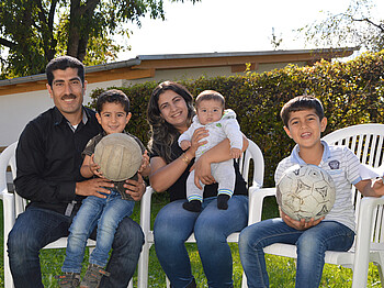 Eine junge Familie mit drei Kindern sitzt in einem Garten auf Plastikstühlen. Sie wirken glücklich und zwei der jungen Buben halten einen Fußball in der Hand. 