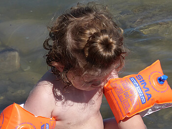 Ein kleines Kind mit orangen Schwimmflügeln kühlt sich im seichten Wasser ab. Die braunen Haare hat es mit einem Dutt nach oben zusammengebunden.  