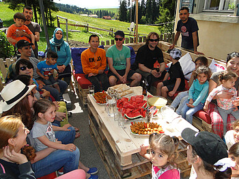 Viele Menschen sitzen im Freien auf selbstgemachten Holzpaletten Sofas. in der Mitte befindet sich ein Holztisch mit verschiedenen Häppchen.