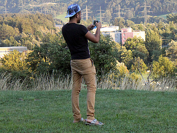 Ein junger Flüchtling steht mit dem Rücken zur Kamera in einer Wiese. Er hält sein Handy in die Luft und es scheint, als würde er ein Foto machen. Er trägt eine beige Hose, ein schwarzes T-shirt und eine Kappe.