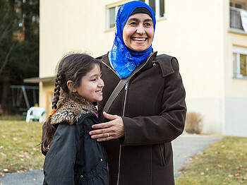 Zwei Flüchtlinge aus dem Nahen bzw. Mittleren Osten, eine Mutter mit ihrer kleinen Tochter, stehen vor einem Flüchtlingsheim. Sie sind beide schlicht und warm bekleidet. Die Mutter trägt neben einer dicken Jacke auch eine blaue Kopfbedeckung. Beide lachen dabei und sind glücklich. 