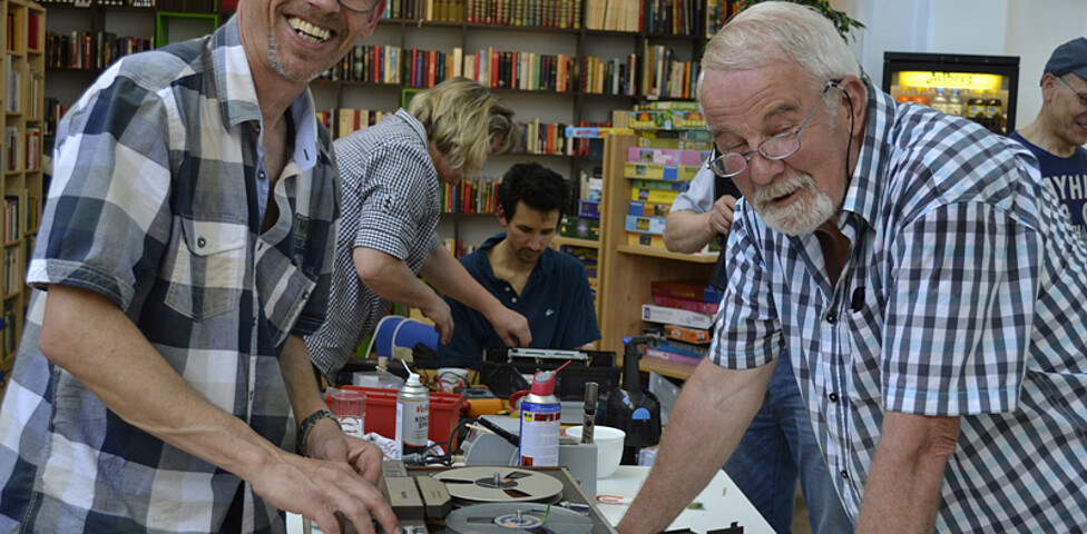 Ein älterer Herr und Mann mittleren Alters, die beide karierte Hemden tragen, reparieren ein altes Aufnahmegerät, das auf einem weißen Tisch steht. Die Stimmung ist entspannt und beide lachen. 