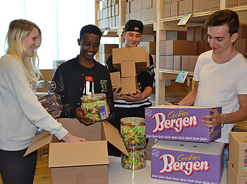 Vier junge Menschen packen Süßigkeiten in Kartons. Sie befinden sich in einem Lager. Alle strahlen.