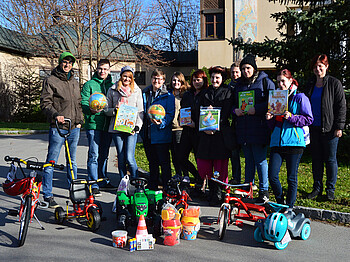 Eine Gruppe junger Erwachsener steht draußen vor einem Wohnblock und zeigt stolz Spielzeug, das die Kinder der MUKI bekommen. Darunter befinden sich Spielzeugtraktoren und Fahrräder mit Stützrädern. Auch Bücher sind dabei. 