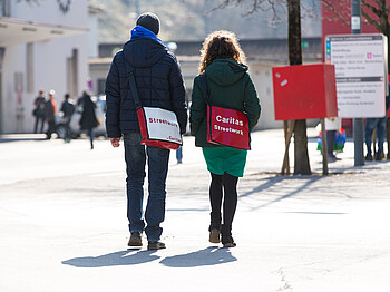 Zwei junge Erwachsene, eine Frau und ein Mann, spazieren Richtung Bahnhof Feldkirch. Sie wurden von hinten fotografiert. Beide tragen sie ein Umhängtasche auf denen jeweils "Streetwork" und "Caritas" steht. Beide sind sie in Winterkleidung angezogen. 