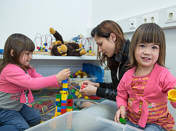 Eine junge Frau spielt mit zwei kleinen Mädchen am Boden Lego. Ein Mädchen im Vordergrund wirkt sehr fröhlich und lächelt. Im Hintergrund baut die junge Frau mit dem Mädchen einen Turm.