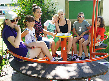 Vier Kinder und drei erwachsene Frauen sitzen in einem orangen Karusell auf einem Spielplatz. Die Kinder, die ca. sechs Jahre alt sind, sind durch das viele Spielen bereits müde und schlaff. Eines lehnt sich an eine Frau an. 