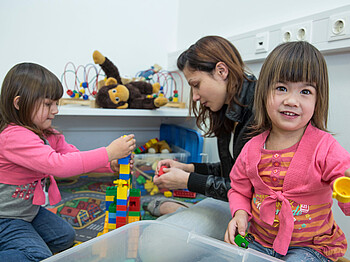 Eine junge Frau mit braunen Haaren spielt am Boden mit zwei kleinen Mädchen mit Lego. Die Frau trägt eine schwarze Lederjacke und die Mädchen rosa Oberteile. 