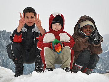 Drei Kinder unterschiedlicher Ethnien spielen im Schnee. Sie tragen dicke Winterbekleidung, die schwarz, weiß, rot und braun ist. Sie sitzen dabei in einer Hocke auf einem Schneehügel. Alle drei Kinder sind erfreut und haben Spaß. Einer der drei Jungs zeigt noch das Victory Zeichen mit beiden Händen. 