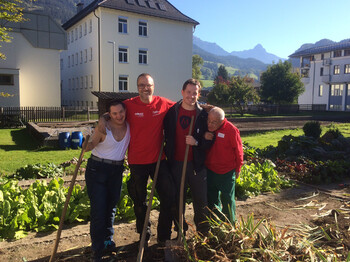 Mitarbeiter von Mondelez in Bludenz helfen im Garten der Werkstätte Bludenz bei schönem Wetter. Zwei Mitarbeiter und zwei Menschen mit Beeinträchtigung stehen im Garten zu einem Gruppenfoto zusammen. 