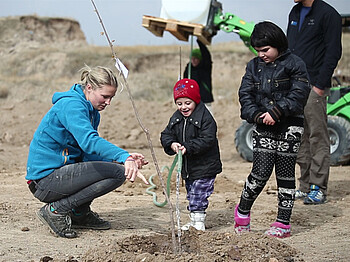 Zwei armenische Kinder planzen mit Hilfe einer Freiwilligen einen Baum in der Landschaft. Alle sind dabei in warmen Winterklamotten gehüllt und dunkel bekleidet. Die junge Frewillige trägt im Kontrast zu den anderen drei sich auf dem Foto befindenen Personen einen hellblauen Sweater. Die Kinder sind angesichts der Situation und der Aufmerksamkeit, die sie erfahren, erfreut. 