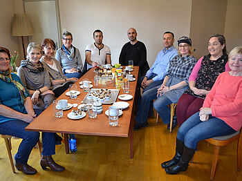 Mehrere Menschen sitzen in einer Runde um eine lange Tafel. Auf dem Tisch befinden sich Snacks und Getränke.