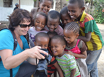 Caritas-Mitarbeiterin Elke Kager zeigt äthiopischen Kindern Fotos auf ihrer Kamera. Die Kinder schauen gespannt und sind erfreut. Elke trägt eine blaue Bluse und die Kinder sind bunt, in unterschiedlichen Farben gekleidet. 