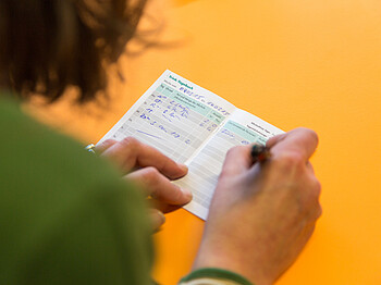 Eine Person mit grünem Oberteil notiert etwas in einem kleinen Heftchen mit der Aufschrift "Trink-Tagebuch".