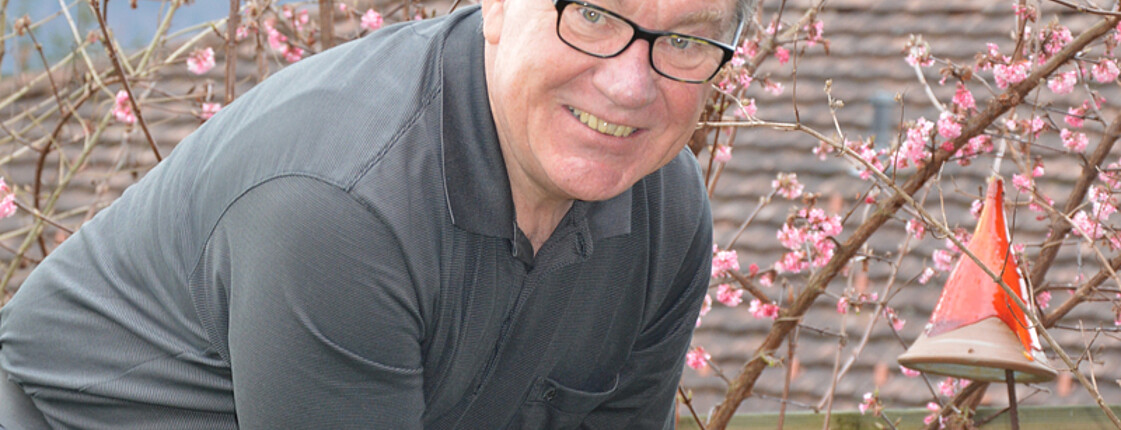 Ein Mann mittleren Alters beugt sich mit den Armen nach unten. Er trägt ein graues Oberteil und eine schwarze Brille. Schauplatz ist im Freien.