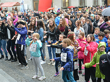 Eine große Ansammlung von Jugendlichen befindet sich in einer Innenstadt. Sie alle nehmen teil an einem Laufwettbewerb. Alle haben sie Wettkampfnummern. 